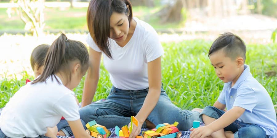 an Asian mom with her three young children building with tegu blocks together  on a picnic blanket in a park. social activities for toddlers