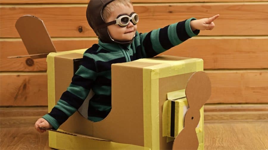 rainy day crafts for toddlers-toddler boy wearing, a striped sweater, a pilot helmet and goggles playing in his cardboard box creation plane.