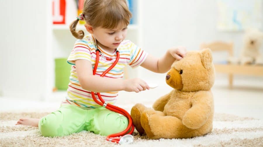 imaginative play-toddler girl on the floor wearing a stethoscope is engaged in pretend play with her bear. she is pretending to feed him medicine with a spoon.