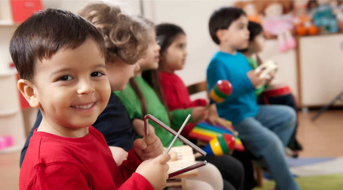 A preschool boy wearing a red shirt plays a triangle in a classroom with other children playing musical instruments. The boy looks at the camera and smiles on Music Monday during the Week of the Young Child. 